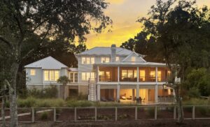 Elevated rear facing photo of coastal home at dusk with lots of windows, huge screened porch, and outdoor entertaining space at ground level.