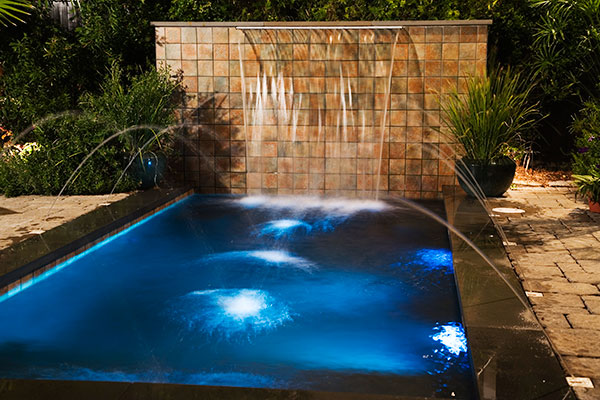 Illuminated pool at night with side water jets and waterfall feature at end.