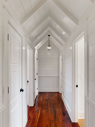 Hallway with horizontal shiplap on walls above vertical beadboard wainscot. Ceiling is vaulted with applied painted wood and intermittent beams spaced down the hall.